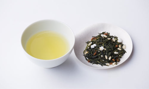 White teacup of light Genmaicha green tea with roasted rice beside small plate of dried bancha tea leaves with roasted rice