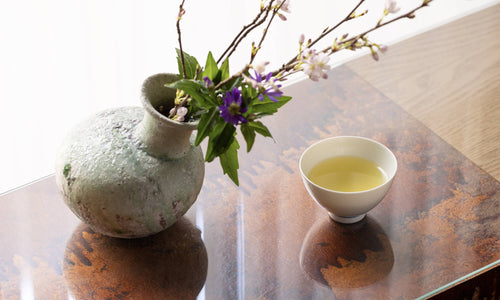 Small porcelain teacup containing fresh green tea beside asymmetrical stone vase with blooming branch of cherry blossom tree