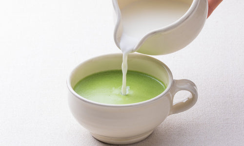 Pouring foamy frothed milk from silver pitcher into concentrated green matcha tea latte base in short round beige ceramic mug