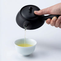 Hand pouring green tea from a black Tokoname-yaki kyusu teapot with side handle Ippodo Tea, filling a white teacup.