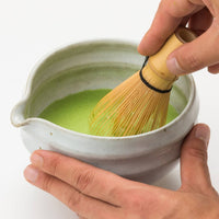 Holding white tea bowl with spout whisking thin usucha Ippodo Tea Shoin matcha with Chasen bamboo 80-tip whisk white table