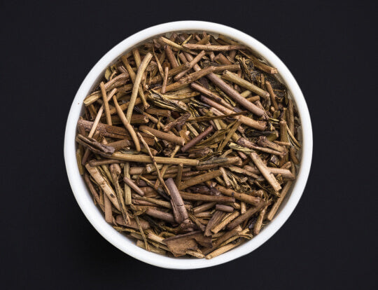 Loose dried brown Ippodo Tea Co. Stems (Kuki) Hojicha Japanese roasted green tea stems in white bowl on black background