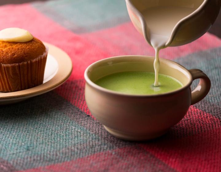 Pouring milk from creamer into matcha tea latte in wide short cream mug beside cupcake on small plate on checkered tablecloth