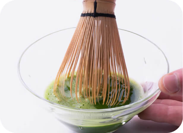 Whisking Japanese matcha green tea in glass bowl on white table with 80-tip bamboo chasen whisk by Ippodo Tea