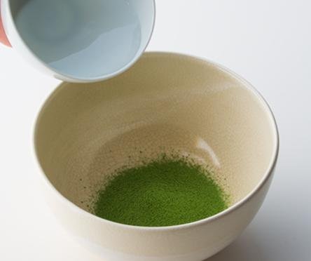 Pouring hot water from white porcelain teacup into cream ceramic tea bowl containing vibrant green sifted matcha