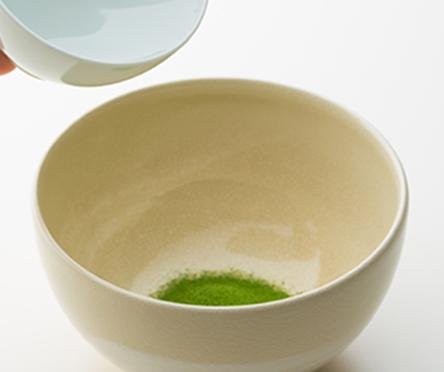 Pouring hot water from white porcelain teacup into cream ceramic tea bowl containing vibrant green sifted matcha