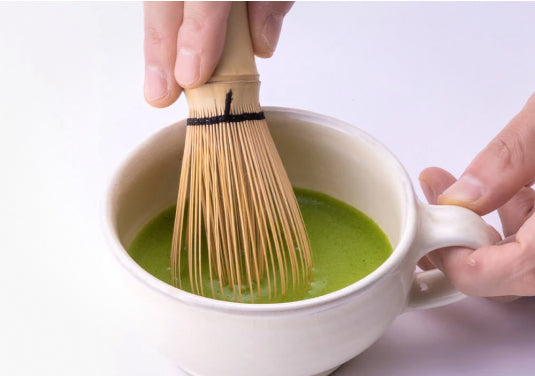 Whisking green matcha base in teacup with handle using Chasen bamboo whisk and overlaid directions arrows along sides of bowl