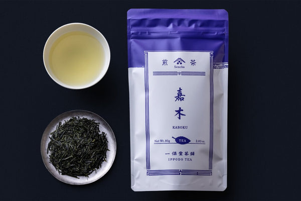 Purple packaging for Ippodo Kaboku Sencha alongside teacup of light green brewed Sencha tea and plate of dried tea leaves