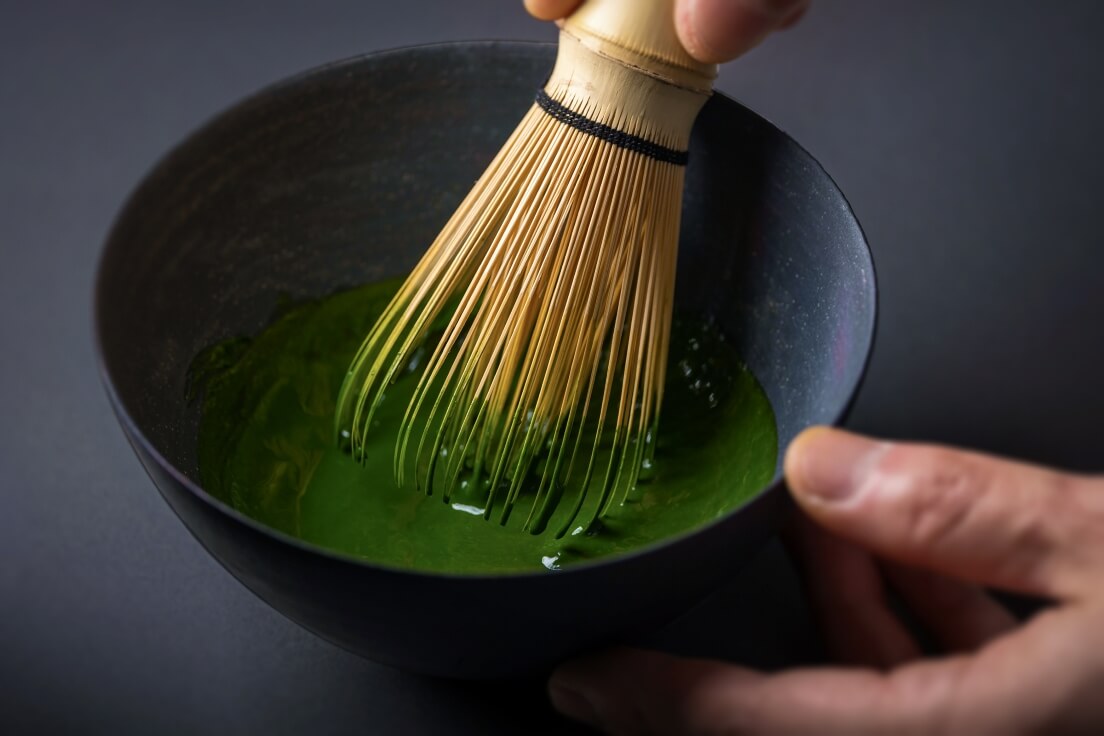 Whisking verdant green koicha matcha in speckled charcoal ceramic tea bowl on dark table using 100-tip Japanese bamboo Chasen
