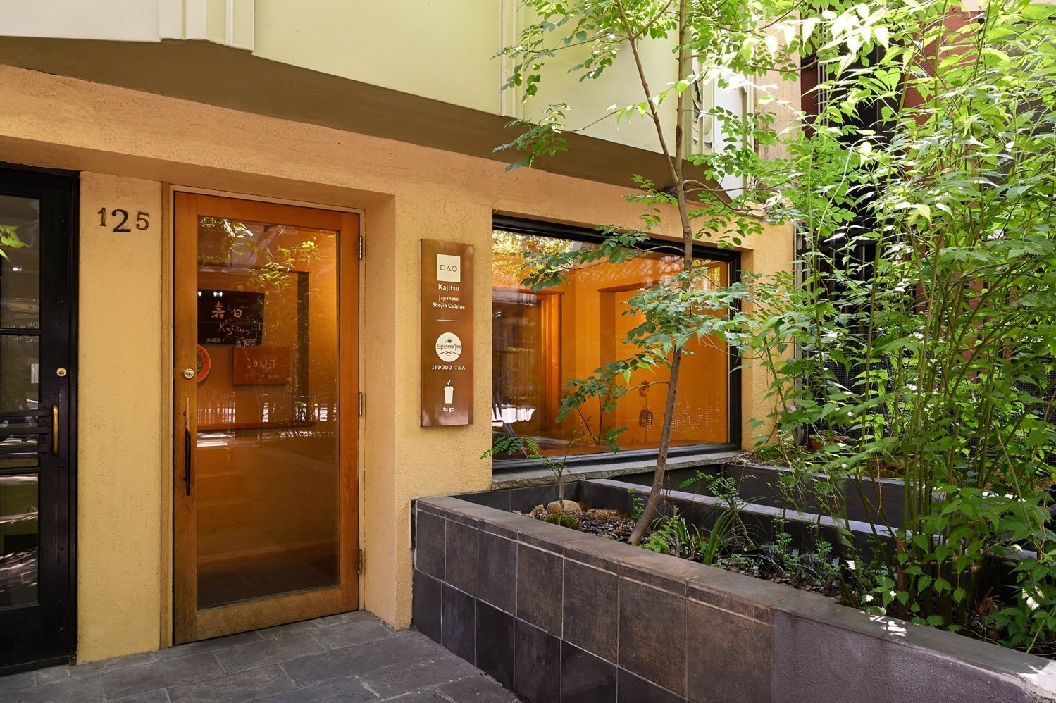 Warmly lit light tan Ippodo Tea storefront in New York City with large window and concrete planter of green foliage ouside