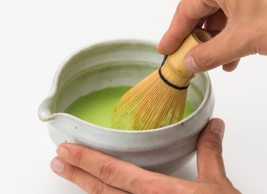 Holding white tea bowl with spout and finger-groove and whisking vivid green foamy matcha tea using Chasen bamboo whisk