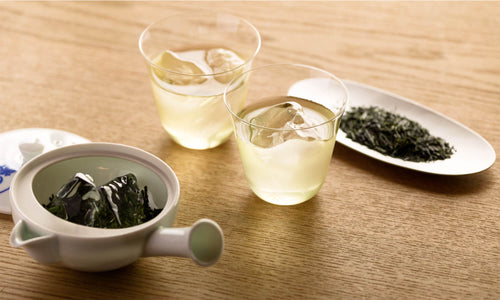 Small porcelain kyusu with lid open showing tea inside beside two glasses of iced light green tea and silver plate of gyokuro