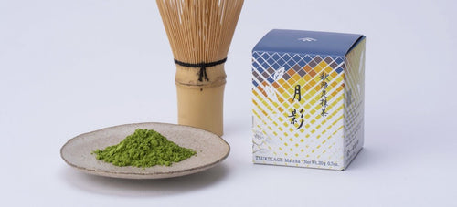 A product photo of Tsukikage fall matcha, showing the box, a bamboo whisk, and a pile of vibrant green matcha powder on a ceramic plate against a white background.