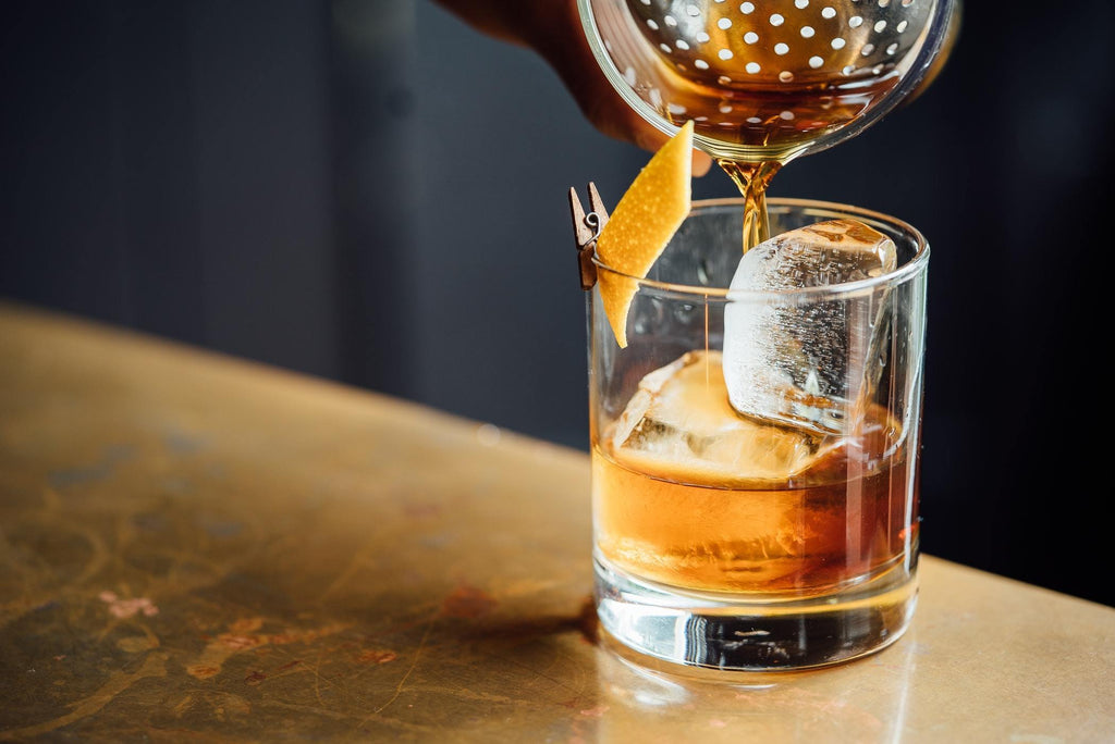 Pouring old fashioned cocktail with orange peel garnished through stainless steel strainer into rocks glass with ice on bar