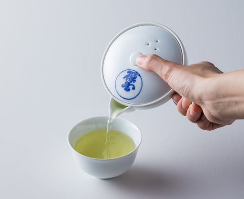 Pouring light green tea from white porcelain Hasami-yaki kyusu teapot with blue logo into white porcelain Japanese teacup