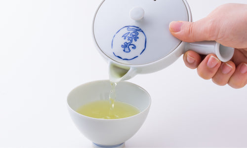 Pouring light green tea from white porcelain Hasami-yaki kyusu teapot with blue logo into white porcelain Japanese teacup