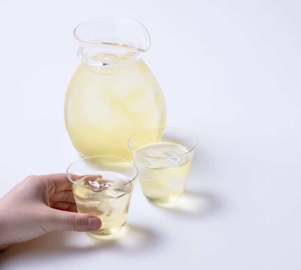 Holding one of two glasses of light yellow Japanese green tea over ice in front of large curved carafe of light colored tea