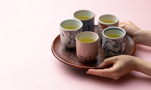 Host holding red brown lacquered wooden Marubon serving tray by Ippodo Tea Co. filled with six decorative cups of green tea