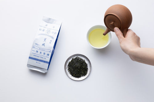 Pouring tea from Yakishime clay kyusu into porcelain teacup beside blue and white package of sencha and plate of tea leaves