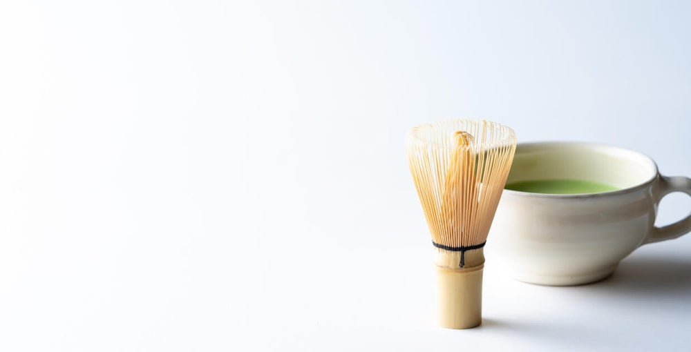 80-tip chasen bamboo whisk sitting upright on end on white table beside beige ceramic tea mug of pastel green matcha latte