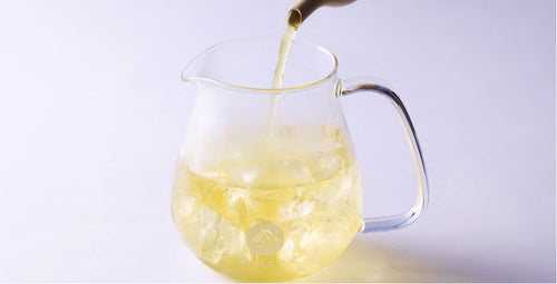 Green tea being poured from elegant spout of ivory teapot into small rounded modern glass pitcher with white Ippodo Tea logo