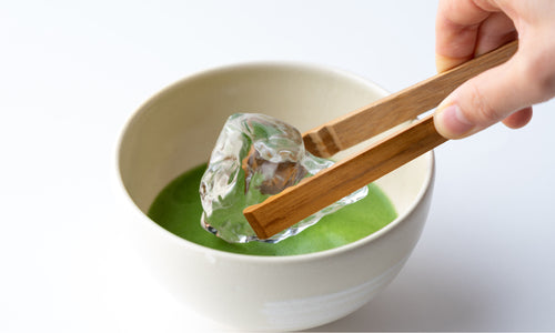 Holding clear ice cub with bamboo tongs and placing it in vibrant green matcha tea base in cream ceramic Japanese teacup