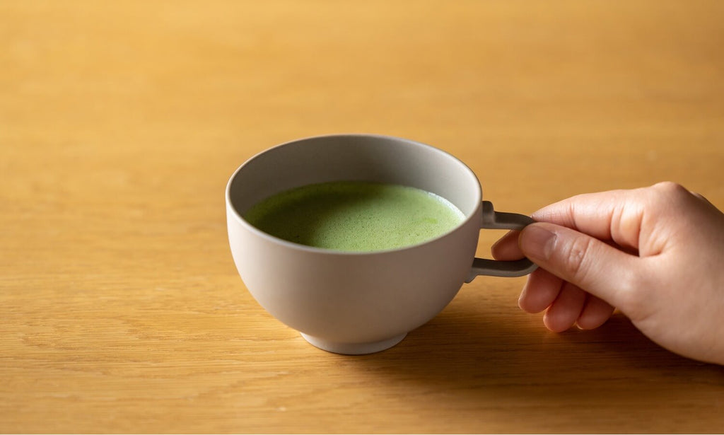 Holding handle of sleek modern gray bowl-shaped ceramic teacup of foamy green Ippodo Tea matcha on wooden table