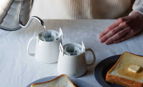 Person pouring water from silver teapot through clipped Drip Teabags into two white mugs with plates of butter toast on table