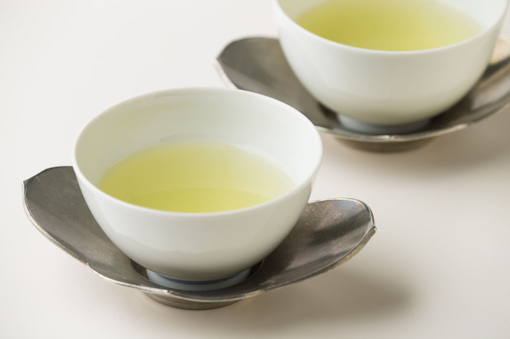 Two small white porcelain Japanese tea cups of light color shincha green tea on silver metal dual petal form saucers