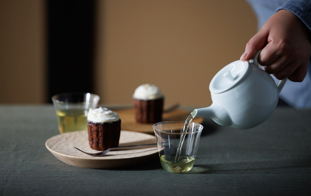 Green tea being poured into glass from white porcelain Hakuji Kyusu teapot beside plates of chocolate cupcakes whipped cream