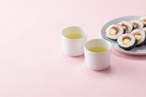 Two modern white handle-less cups of brewed green tea beside grey plate of prepared tofu and mushroom sushi set on pink table