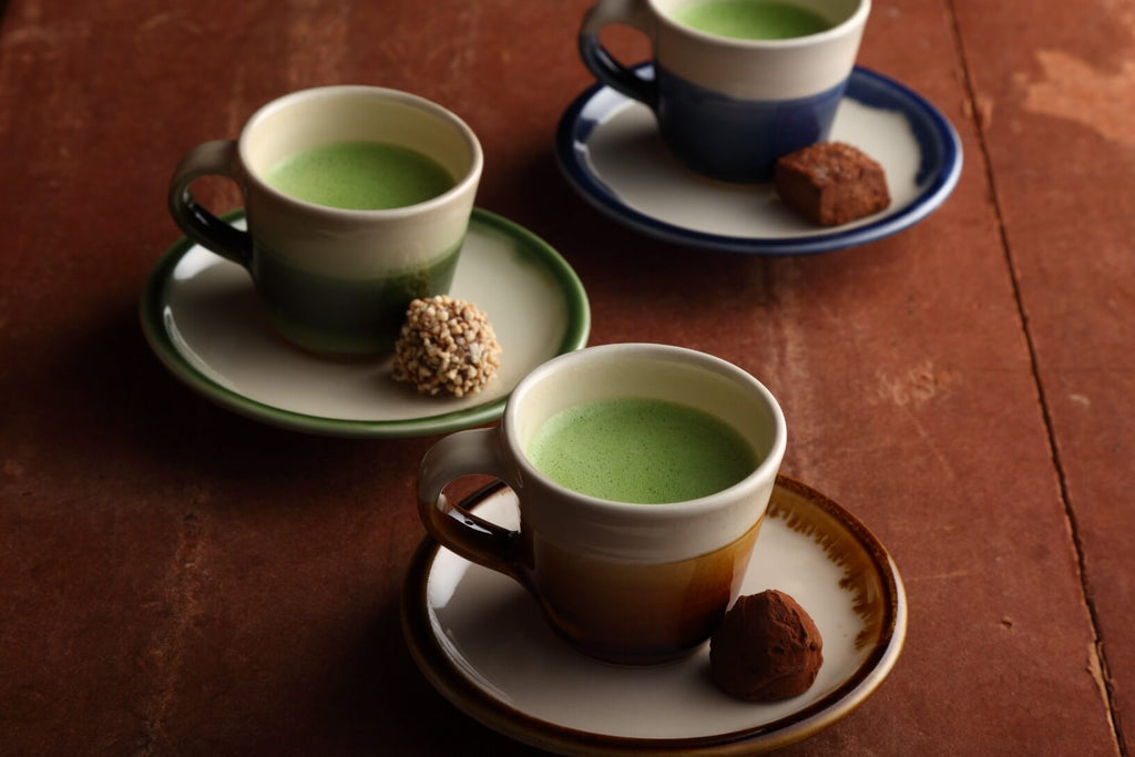 Chocolate desserts on saucers of three different colored teacups containing frothy green Ippodo matcha set on wooden table