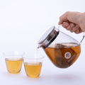 A hand pours Mugicha tea from a clear glass teapot with a metal strainer into two small clear cups on a clean white background. The teapot has a visible circular logo reading IPPOOTEA. - thumbnail 4
