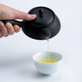 Hand pouring green tea from a black Tokoname-yaki kyusu teapot with left side handle Ippodo Tea, filling a white teacup.