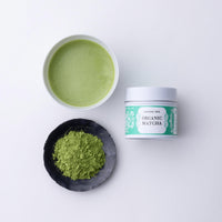 Teacup of green prepared matcha tea beside black plate of loose tea powder and green and white tin of Ippodo Organic Matcha
