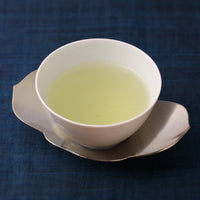 Small white porcelain cup of light color Japanese gyokuro green tea on silver dual petal leaf shaped saucer on navy table