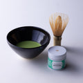 Tin of New Harvest Matcha with green label beside dark tea bowl of prepared matcha by Ippodo Tea and bamboo whisk (Chasen)