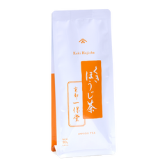 A tall white pouch of Ippodo Tea Stems Hojicha (80g / 2.82 oz) featuring a bright orange vertical banner with Japanese calligraphic text for 