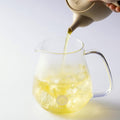 Ippodo Tea; Kanro, being poured from a beige teapot into a glass teapot with a light background