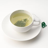 Rounded white mug on matching saucer with Ippodo Tea painted on inner lip holding brewed light green tea and gyokuro teabag