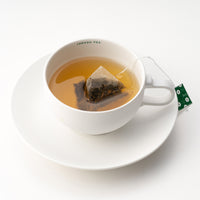 Rounded white mug on matching saucer with Ippodo Tea painted on inner lip holding brewed roasted green tea and hojicha teabag