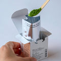 Scooping matcha powder with bamboo tea ladle from foil package inside box of Kan matcha with instructions on back showing