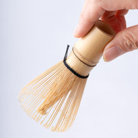 Holding 100-tip bamboo Chasen matcha whisk tea utensil in front of white table