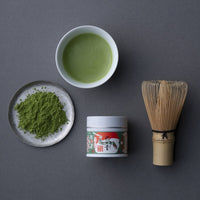 Ippodo's finely-ground green New Year's Matcha loose on silver plate beside bowl of matcha, decorative tin and bamboo whisk