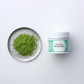 Matcha powder on a silver plate next to a blue-green container labeled 'New Harvest Matcha' on a white background