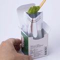 A tea ladle scoops bright green Ippodo Tea Koma-Mukashi (New Year's Matcha 2026) from inner foil pouch of green product box.