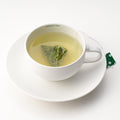 Rounded white mug on matching saucer with Ippodo Tea painted on inner lip holding brewed green tea and sencha teabag