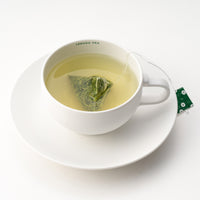 Rounded white mug on matching saucer with Ippodo Tea painted on inner lip holding brewed green tea and sencha teabag