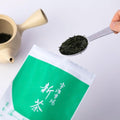 Person scooping tablespoon of New Harvest tea leaves from bright green bag of Uji Shincha tea beside Ivory Kyusu teapot