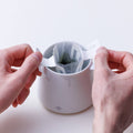 Two hands spreading two panels at top of Drip Teabag across edges of white Ippodo Tea logo mug and clipping teabag to sides